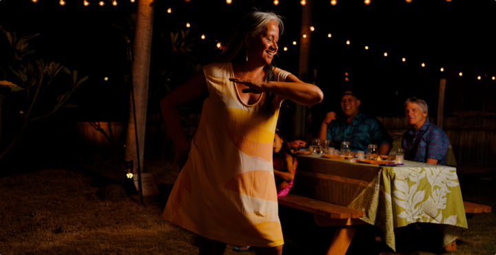 Een lachende vrouw danst de hoela onder sfeervolle lichtjes. Een gezin zit achter haar aan een picknicktafel te eten.