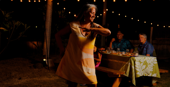 A smiling woman dances hula under string lights. A family sits behind her at a picnic table eating dinner.