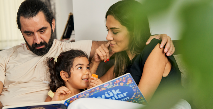 Un hombre, una mujer y una niña leen juntos en un sofá. La niña come una verdura de color naranja mientras mira a la mujer.