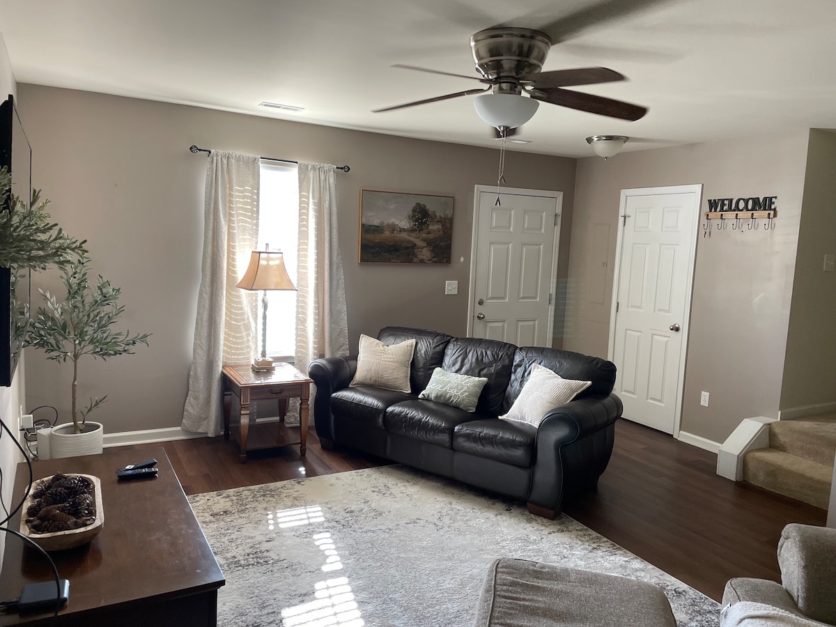 A cozy living room features a dark leather sofa and a light-colored area rug. Natural light filters through sheer curtains, illuminating a wooden side table with a lamp. A potted plant adds a touch of greenery, and a welcoming sign is visible on the wall.