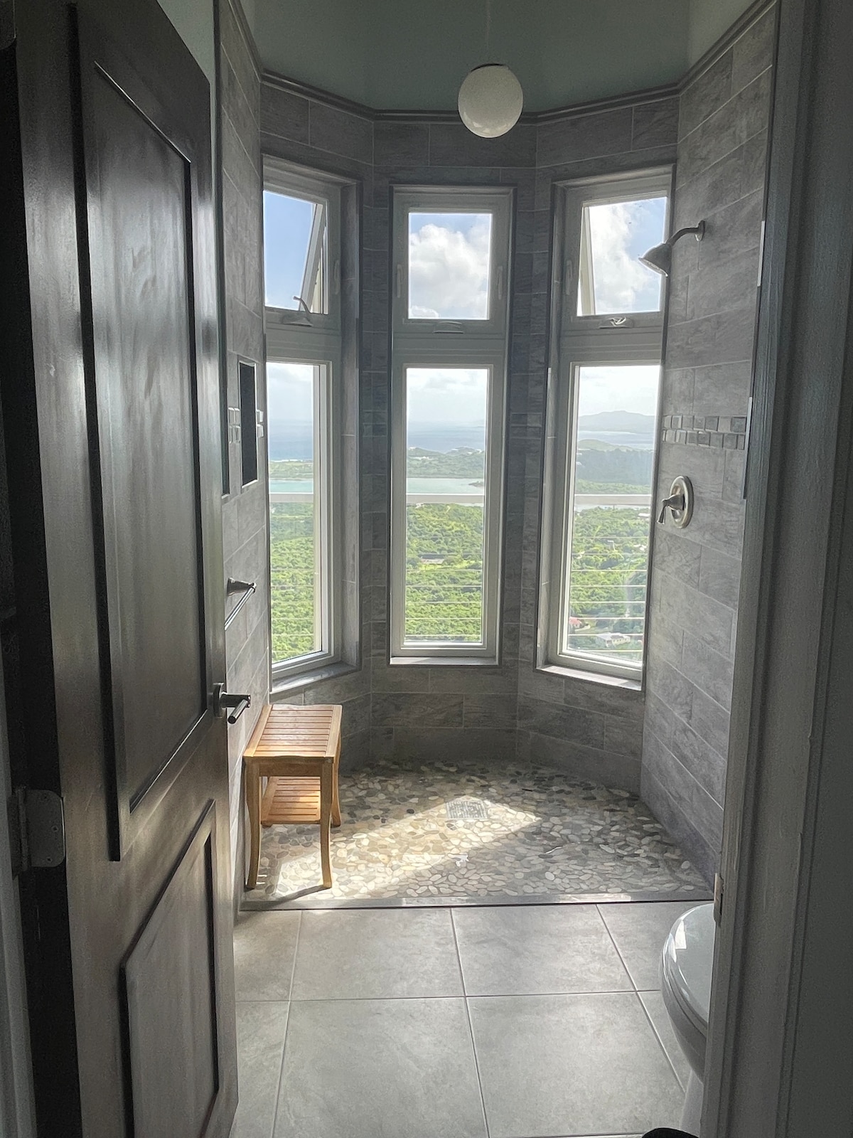 A modern bathroom is showcased with large, clear windows offering scenic views of the landscape. Natural light fills the space, highlighting the stone shower floor and tiled walls. A wooden bench is positioned near the entrance, adding functionality to the serene setting.