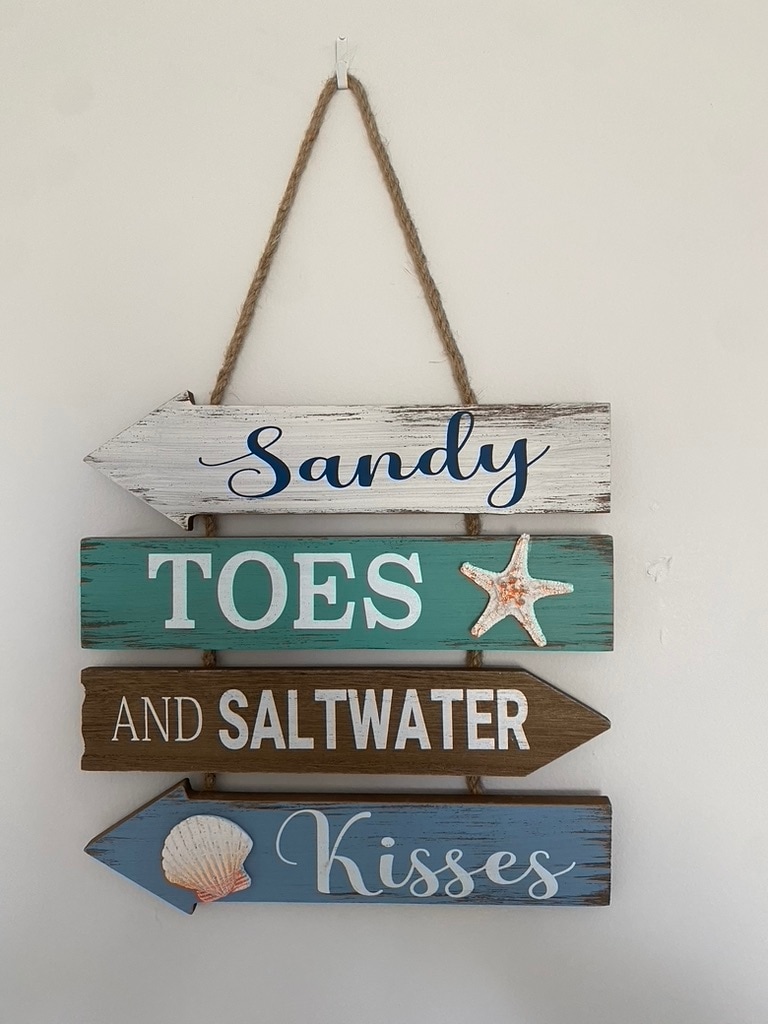 A decorative wooden sign featuring four directional arrows. The words 'Sandy', 'Toes', 'And Saltwater', and 'Kisses' are painted in various colors. A starfish is displayed on the 'Saltwater' arrow, and a seashell decorates the 'Kisses' arrow, adding a coastal theme.