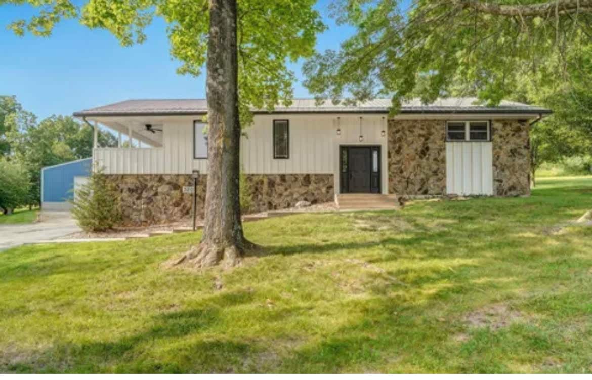 A country-style home is set on a grassy lot, featuring a combination of stone and white siding. The front porch leads to a dark front door, flanked by large windows that allow natural light. Mature trees provide shade in the surrounding area.