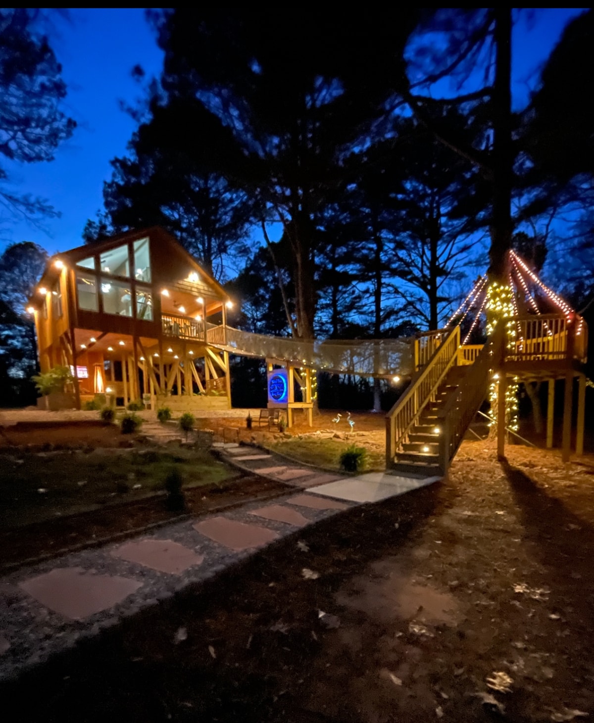 A charming treehouse, lit with warm lights, stands gracefully among tall trees during twilight. A wooden walkway connects the structure to a brightly adorned area planted with shrubs, enhancing the serene ambiance of the surroundings.