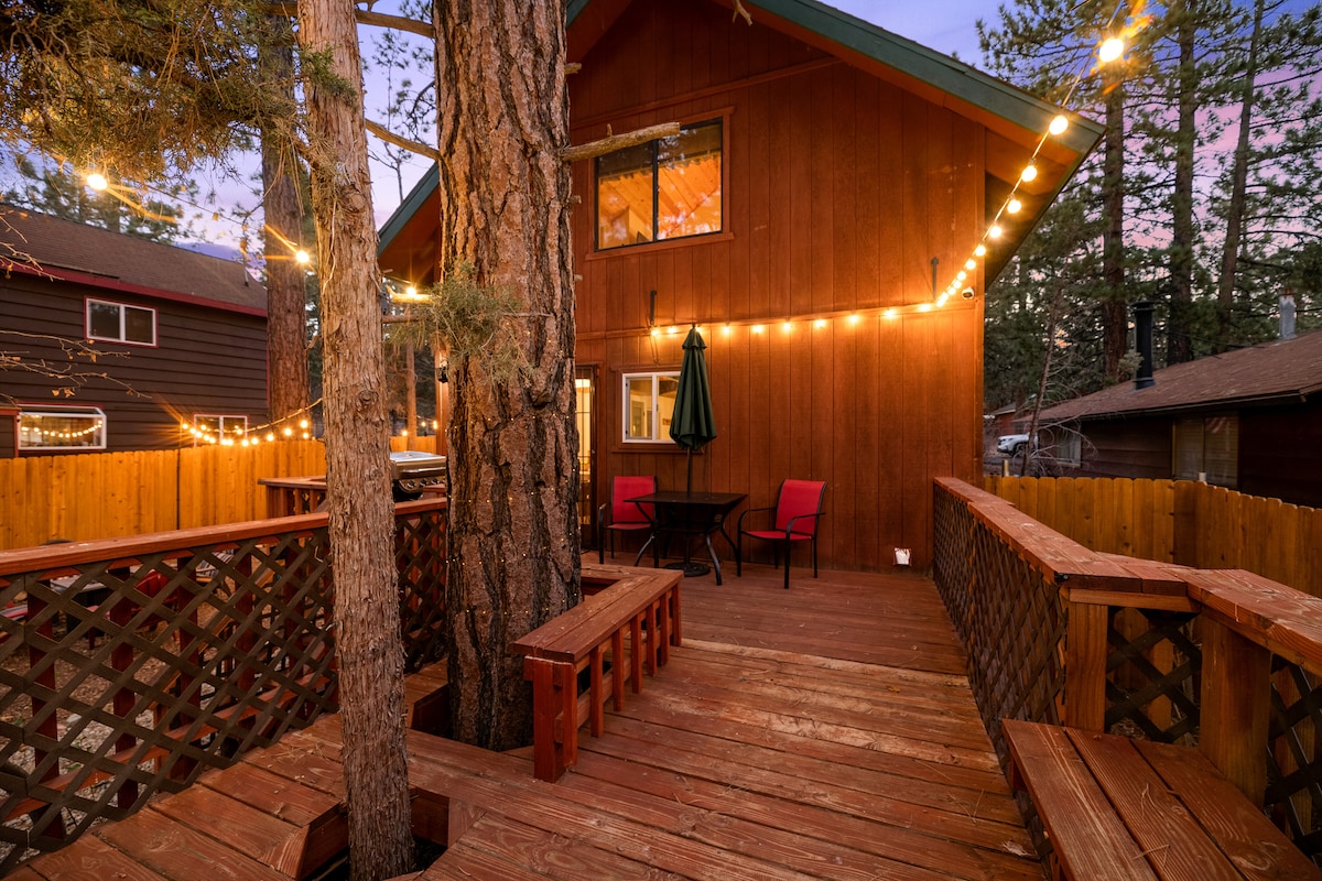 A wooden deck area is visible, framed by tall trees and illuminated by string lights. There are two seating benches and a small table with chairs, creating a cozy outdoor space that invites relaxation amidst a natural setting.