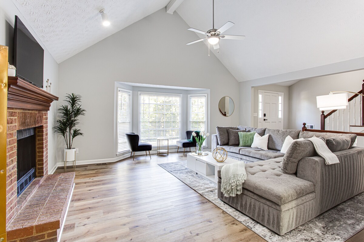 A welcoming living area showcases a spacious sectional sofa, complemented by decorative pillows. A large window bathes the room in natural light, while a flat-screen TV is mounted above a brick fireplace. Two accent chairs are positioned near a modern coffee table.