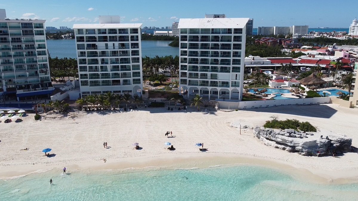 Two multi-story buildings are positioned next to each other along a sandy beach. The coastline features small umbrellas scattered across the shore, with gentle waves lapping at the water's edge. A variety of palm trees provide greenery in the foreground, enhancing the beach atmosphere.