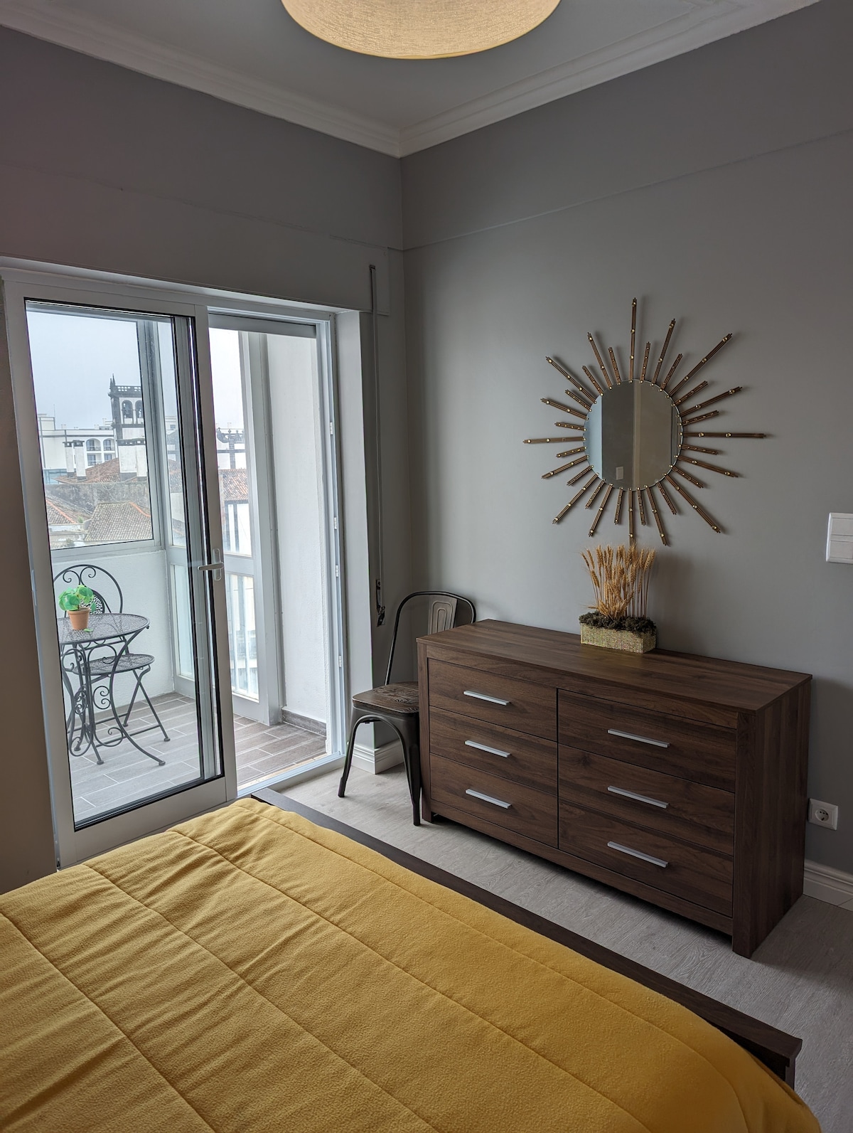 A bright bedroom features a spacious bed covered with a yellow blanket. A wooden dresser stands against the wall, complemented by a decorative mirror. A sliding glass door opens to the enclosed balcony, revealing a small table and chair set, inviting natural light into the room.