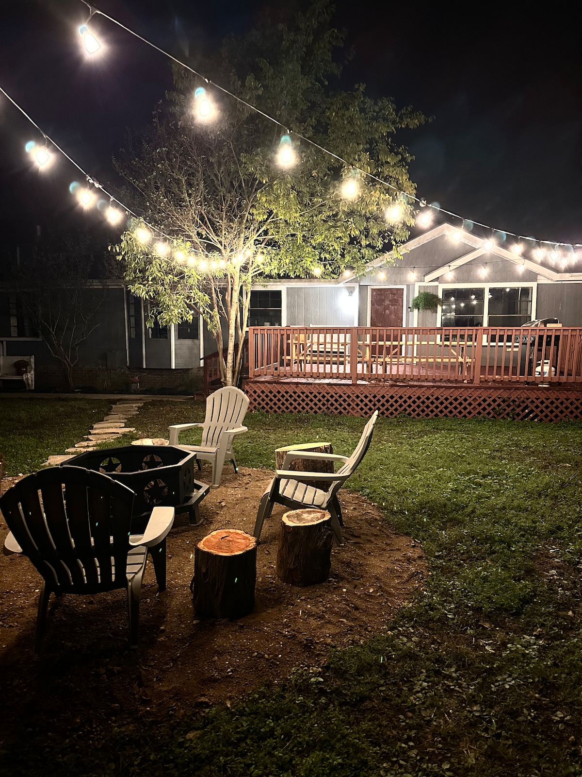 An inviting outdoor space is illuminated by string lights, casting a warm glow over a firepit area. Comfortable adirondack chairs are arranged around the firepit, accompanied by rustic log stools. A well-maintained grassy yard provides a serene backdrop, with a deck visible in the background.