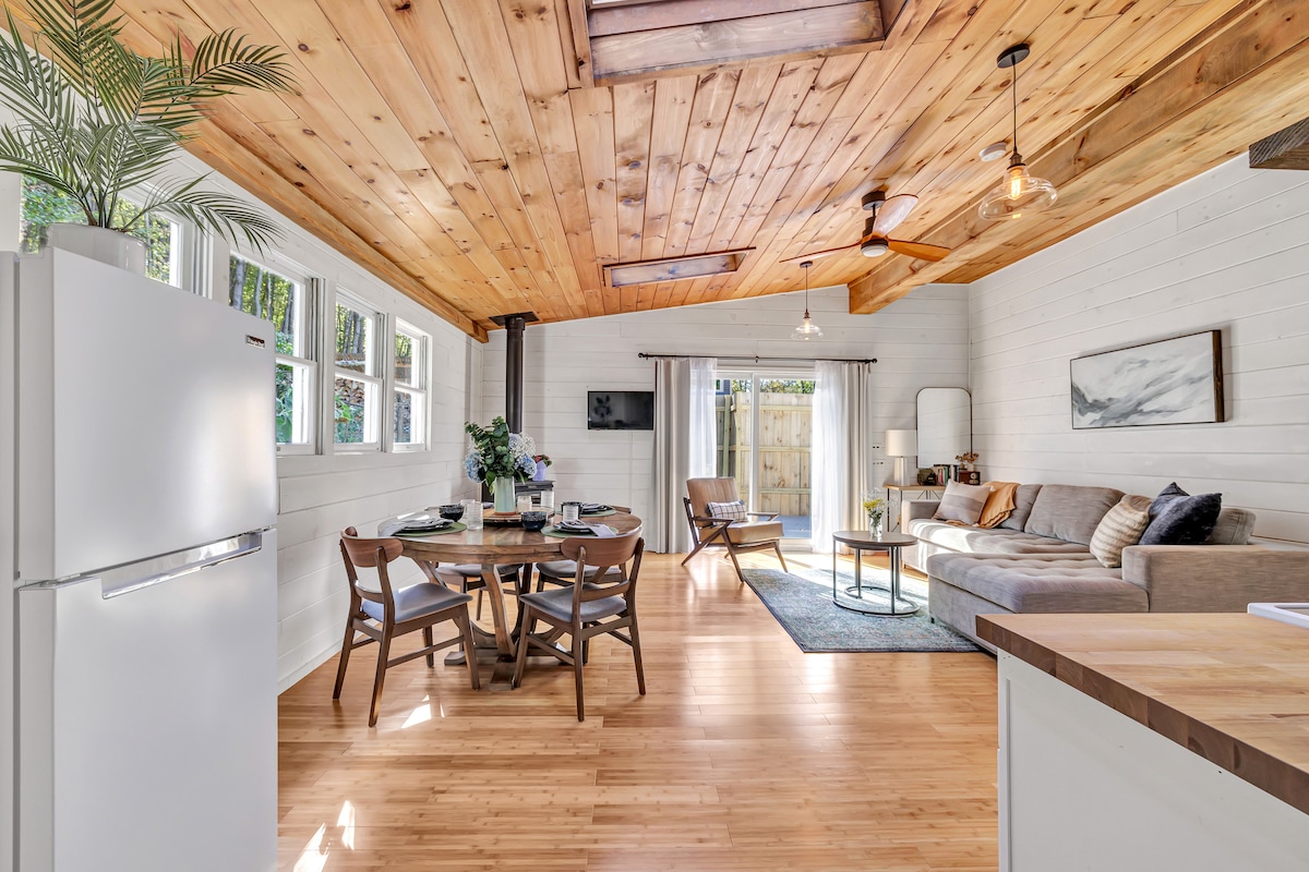 An open-concept living area features wooden ceilings and a bamboo floor. A sectional sofa and coffee table are positioned beside a dining table for four. Sliding glass doors provide access to an outdoor space, while large windows allow natural light to fill the cozy atmosphere.