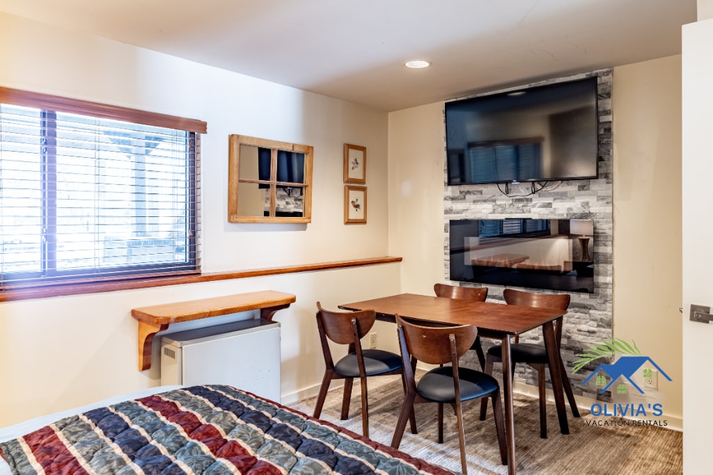 An open living area features a cozy sleeping space and a dining setup. A table with four chairs is positioned next to a modern fireplace, complemented by a wall-mounted television. Natural light filters through a window dressed with blinds.