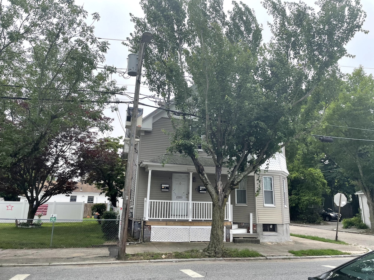 A two-story house is presented from the street view, featuring a welcoming front porch with steps leading up to the entrance. Surrounding trees offer shade, while power lines and street signage can be seen in the vicinity, indicating a residential neighborhood.