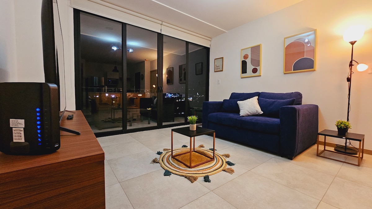 A modern living area features a navy blue sofa positioned beside a small round table. A flat-screen TV is mounted on the wooden console. Large glass doors display city views while providing natural light. Decorative artwork enhances the wall, and a floor lamp illuminates the space.