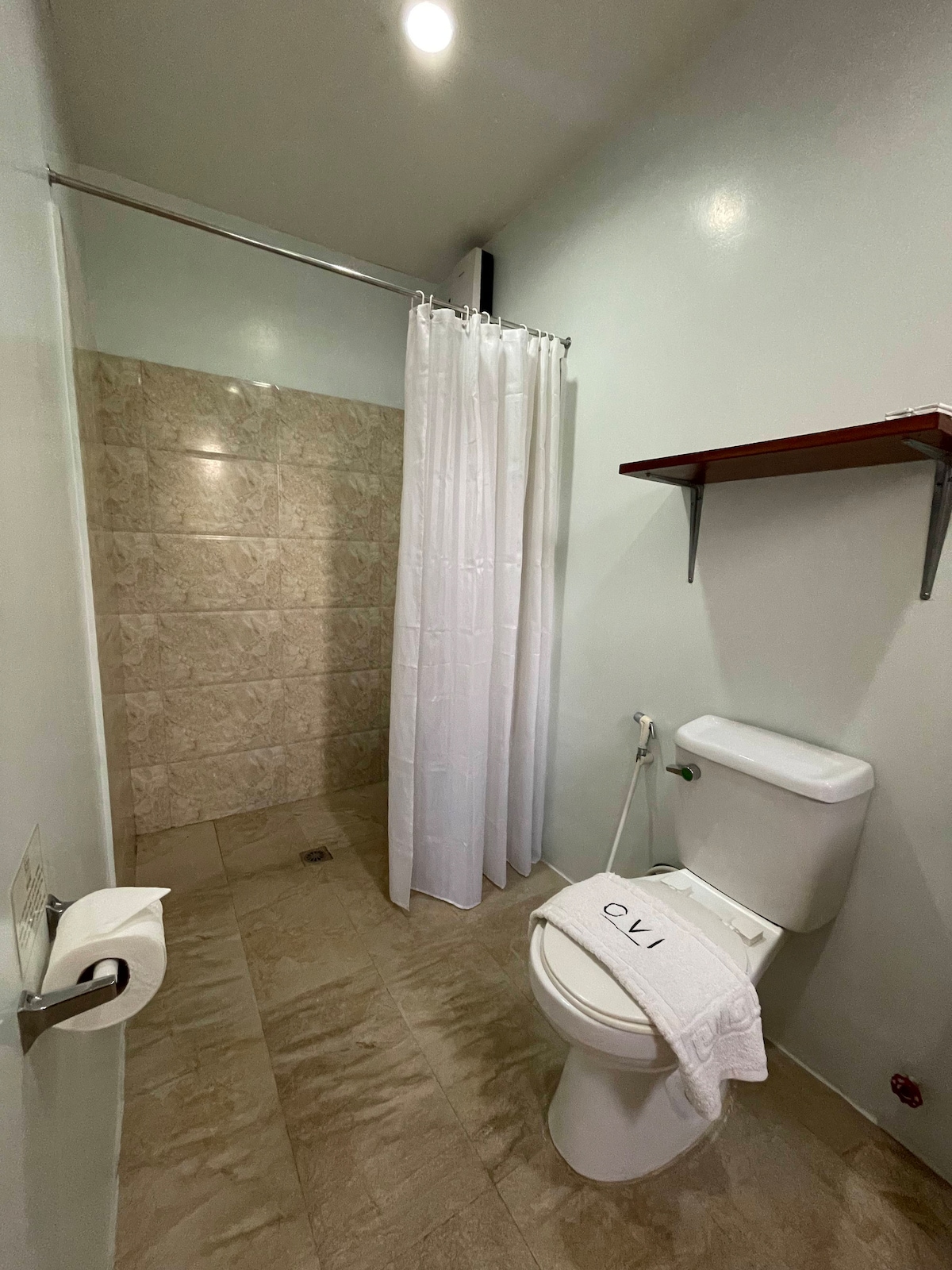 The well-appointed bathroom features a spacious shower area with a translucent curtain and textured wall tiles. A modern toilet is positioned next to a wooden shelf, and a fresh towel is neatly placed on the toilet. Warm lighting enhances the clean, minimalist design.