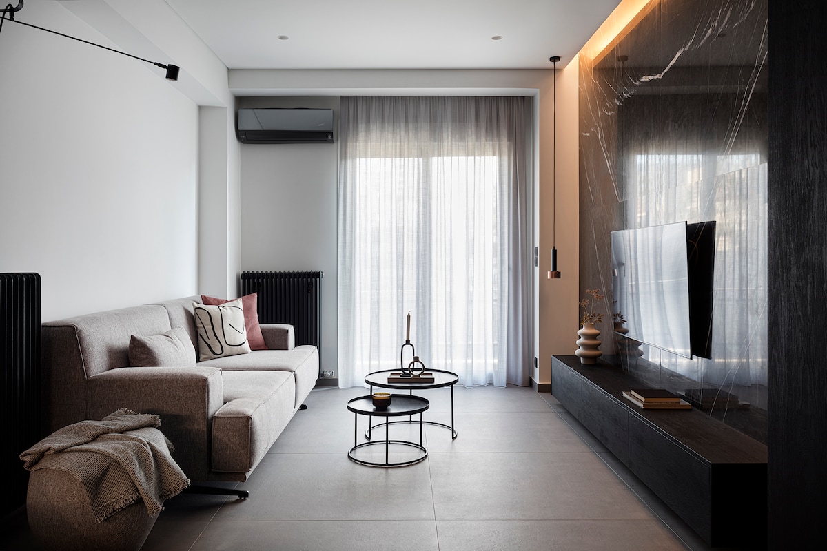 A modern living room is depicted, featuring a gray sofa and a sleek coffee table. Natural light filters through sheer curtains, illuminating the space. A large TV is mounted on a dark feature wall, complemented by minimalist decor elements, creating an understated elegance.