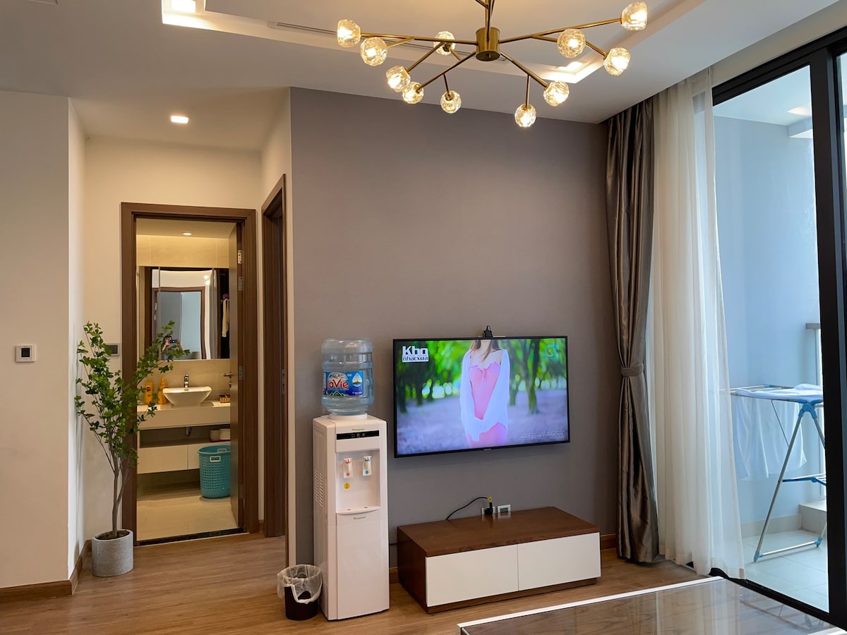 A bright living area features a modern television mounted on a wall above a low wooden cabinet. A water dispenser stands beside the cabinet. Natural light filters through large glass doors leading to a balcony, while tasteful decor elements enhance the simplicity of the space.