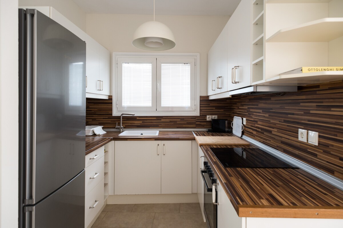 A modern kitchen is displayed with sleek white cabinetry and a wooden countertop. A stainless steel refrigerator and stove are visible, alongside a sink positioned below a window that allows natural light into the space.
