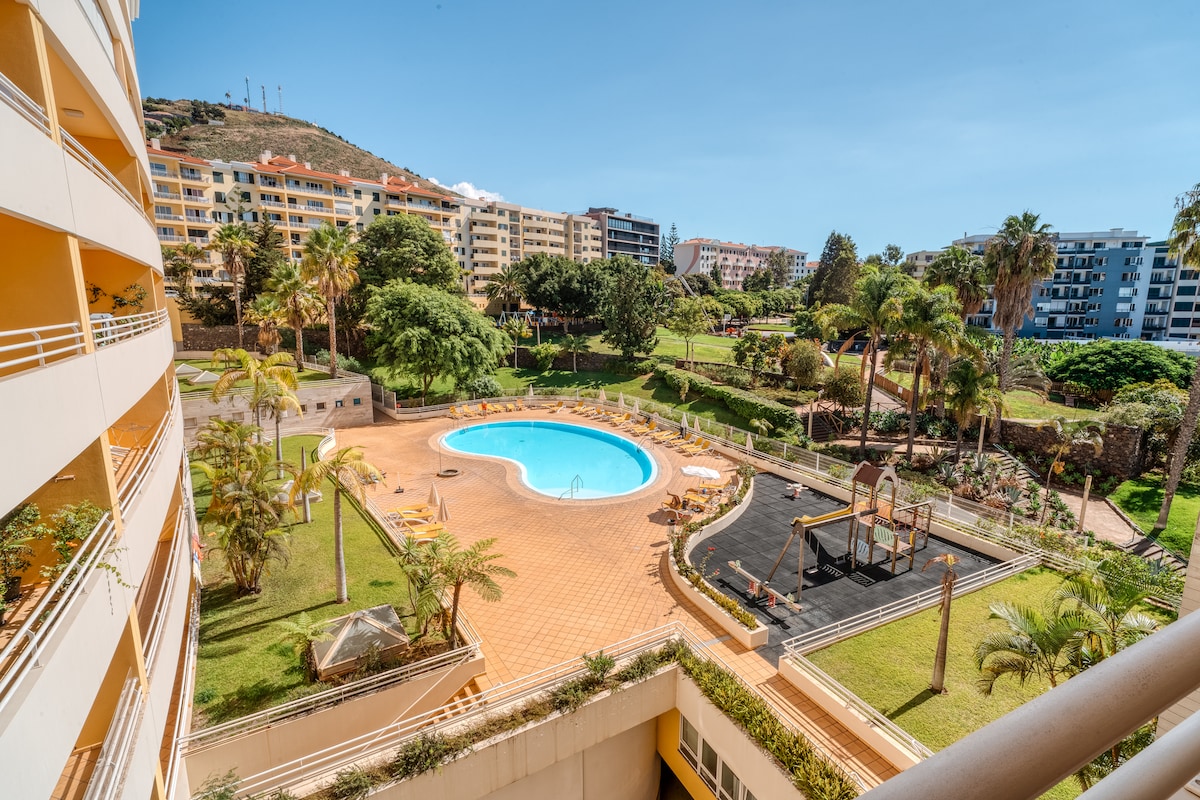 A communal outdoor pool is framed by lush greenery and palm trees. Comfortable lounge chairs are arranged around the pool area, while a playground is visible nearby. The scene captures a sunny day, enhancing the inviting atmosphere of the landscaped grounds.