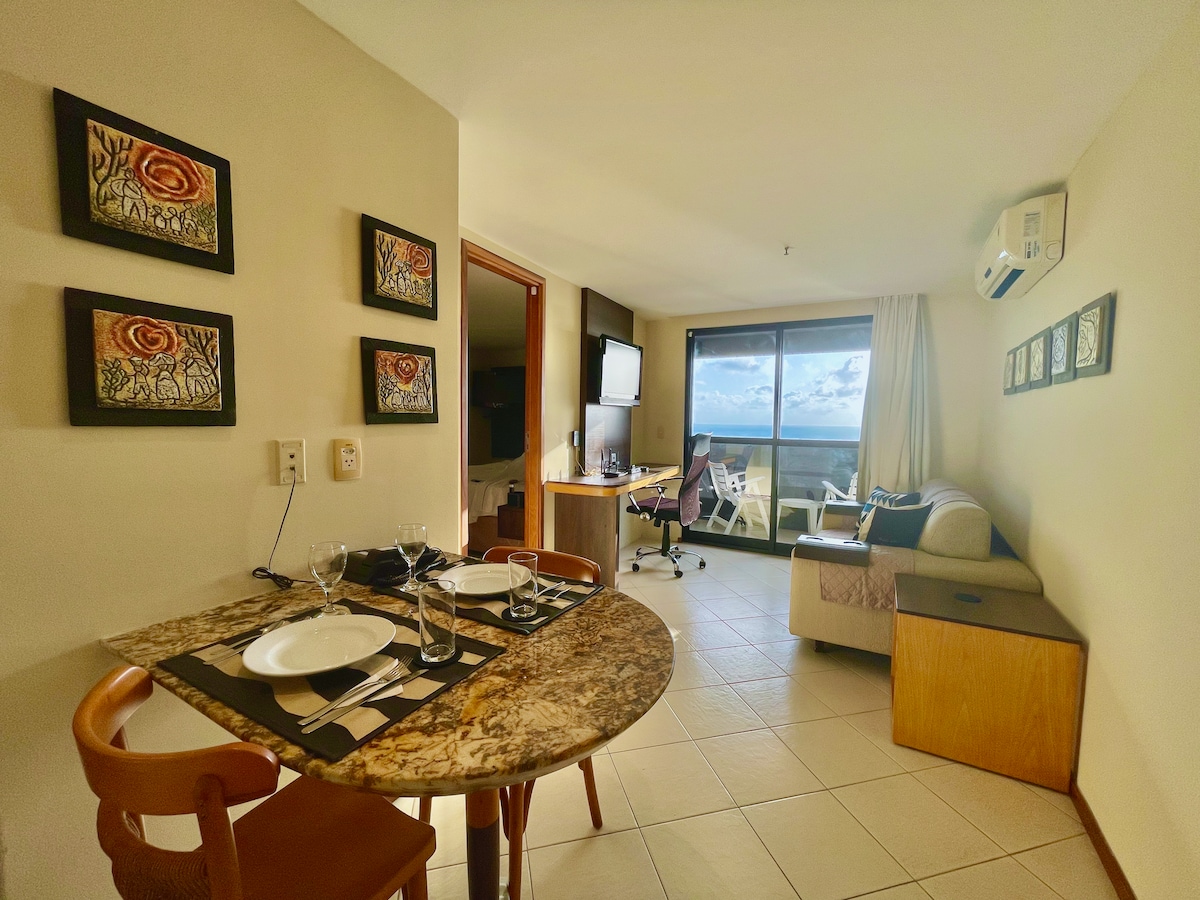 The apartment features an open living space with a dining area equipped with a round table set for two. A comfortable sofa is positioned near a large window with a view, complemented by a work desk and chair. Wall art decorates the room.