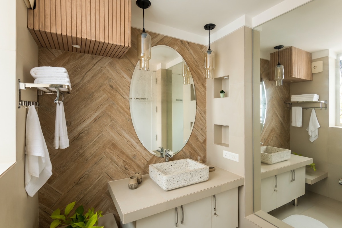 A modern bathroom features natural wood accents and warm lighting. A stylish oval mirror is complemented by hanging pendant lights. A sleek countertop supports a textured basin, while neatly arranged towels and a small plant add subtle touches of comfort.