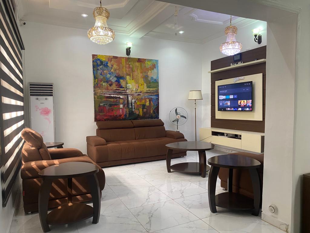 A well-lit living area features a large, colorful abstract painting above a brown sofa. Two side tables and a central coffee table complement the seating arrangement. A standing fan provides airflow, while wall-mounted lights add brightness to the white walls and polished marble floor.