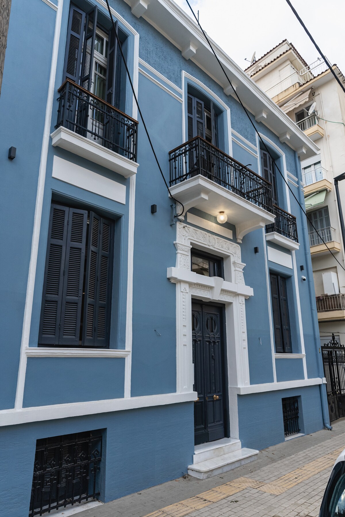 The exterior of a neoclassical building is presented, featuring a blue facade with white trim. Balconies with black railings adorn the upper floors, while symmetrical windows display traditional wooden shutters. A decorative entryway leads to a central door with elegant detailing.