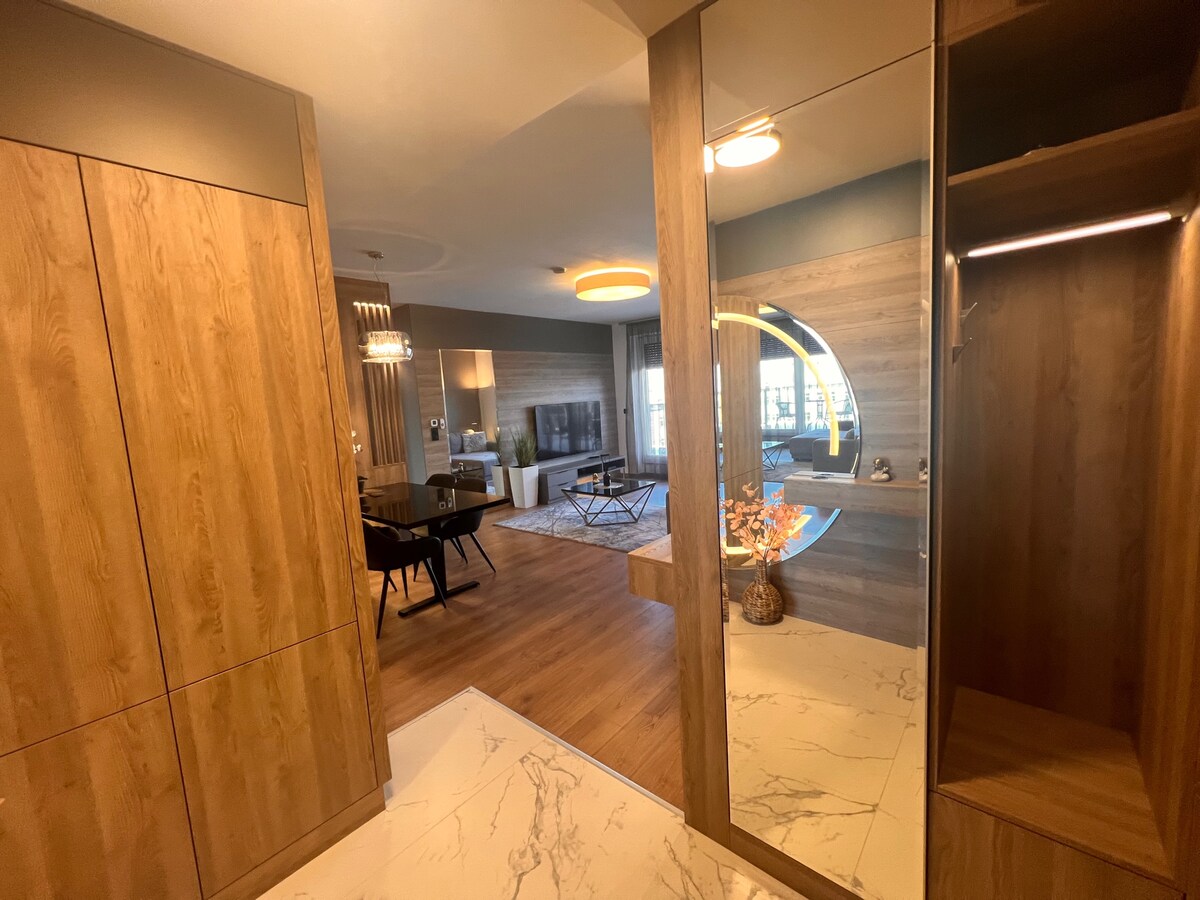 A modern hall is visible, featuring wooden cabinetry and a stylish mirror. The open living area can be seen in the background, showcasing a cozy seating arrangement and a contemporary coffee table. Natural light filters in, enhancing the warm tones of the wooden flooring.