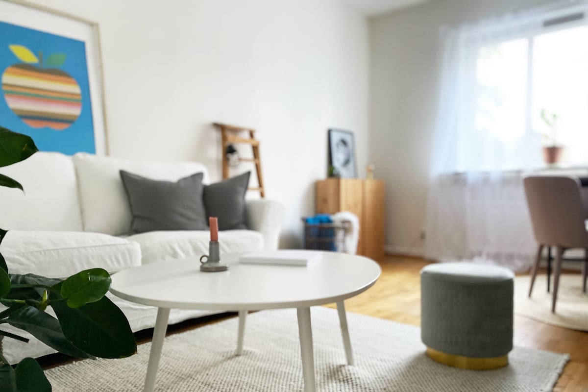 A living area is presented with a white sofa adorned with gray pillows. A round coffee table sits in front of the sofa, accompanied by a textured stool. Natural light filters through sheer curtains, illuminating the space with a relaxed ambiance.