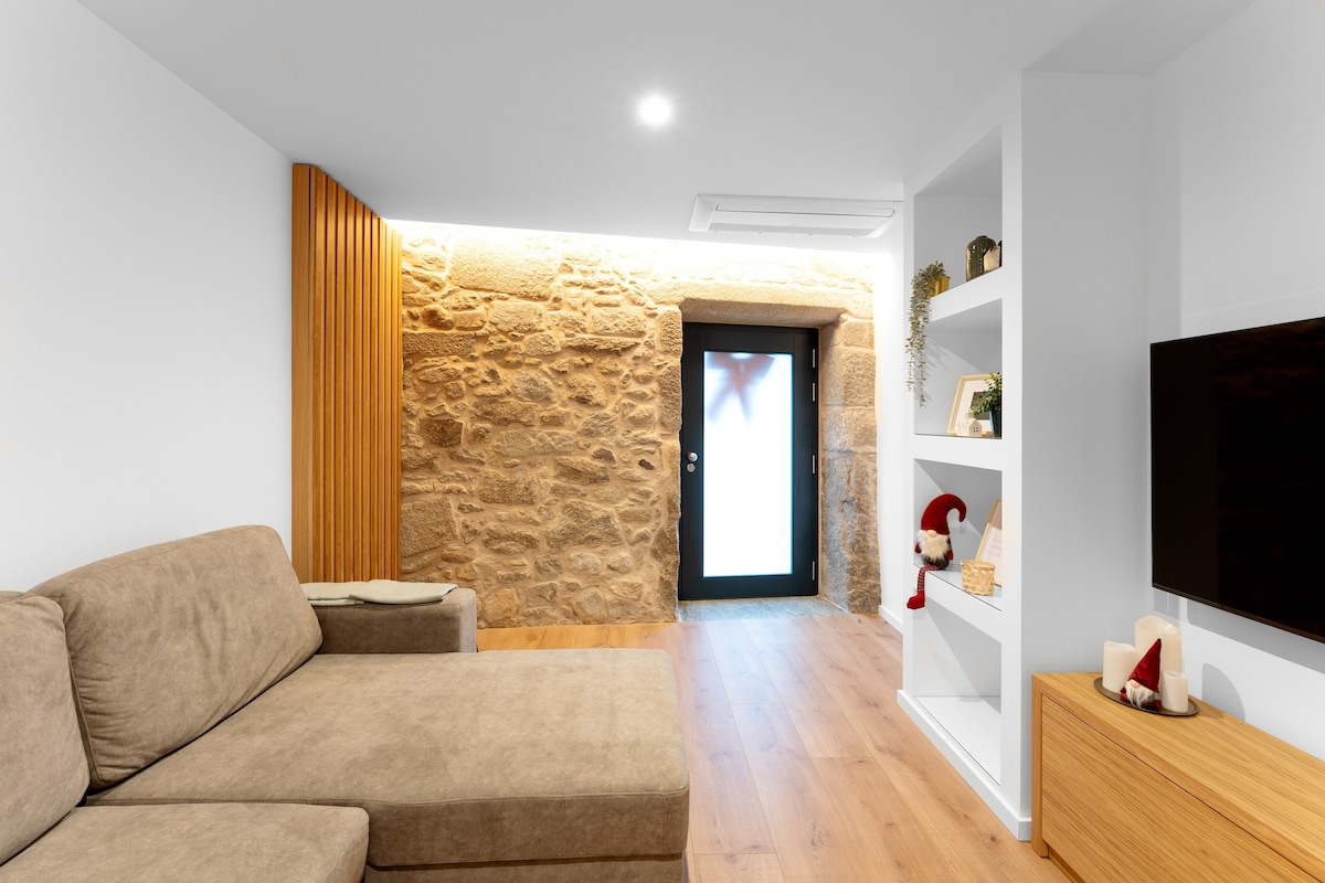 A cozy living area features a soft, beige sectional sofa, complemented by light hardwood flooring. A stone wall adds texture to the space, while a contemporary shelving unit displays decorative items. An entrance with a glass door provides access to the outside.