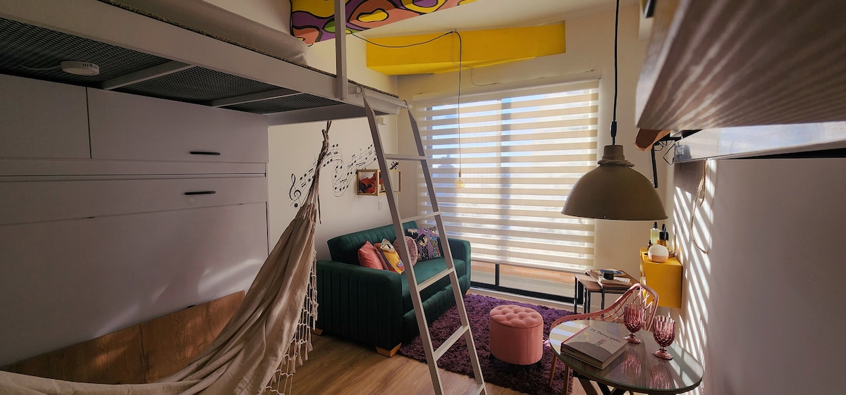 A lofted bed is paired with a cozy seating area featuring a green sofa, a round pink ottoman, and a hammock hanging nearby. Natural light filters through striped window blinds, while warm tones are added by a pendant light and colorful decor on the walls.