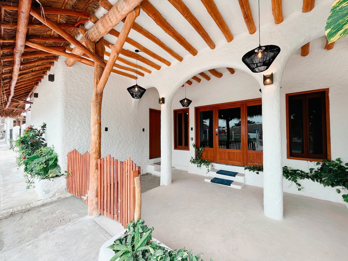 The entrance showcases a charming, open facade with wooden beams and a smooth white exterior. Multiple hanging light fixtures illuminate the space. Decorative plants line the path leading to a spacious doorway with glass windows, inviting natural light indoors.