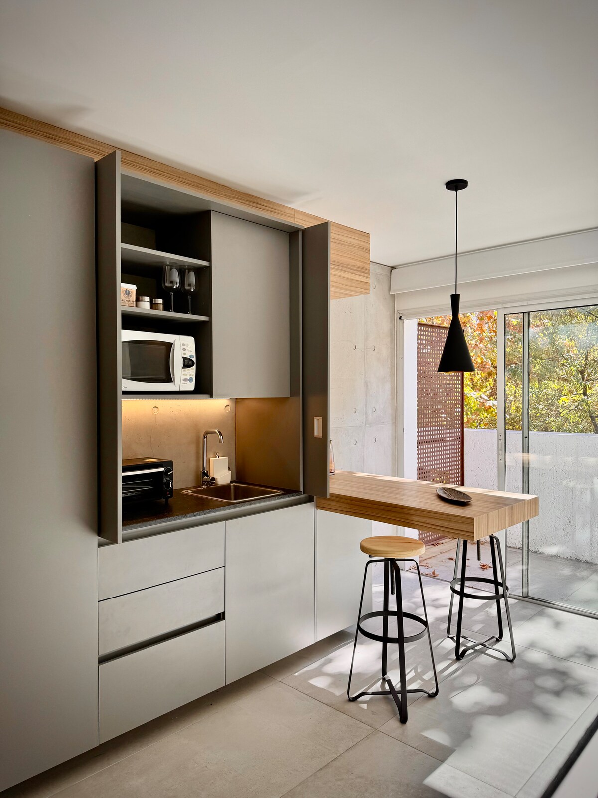 A modern kitchen space features sleek cabinetry with a built-in microwave and sink. Minimalist bar stools are positioned at a light wooden countertop. A sliding glass door allows natural light to enter, revealing a view of greenery outside.