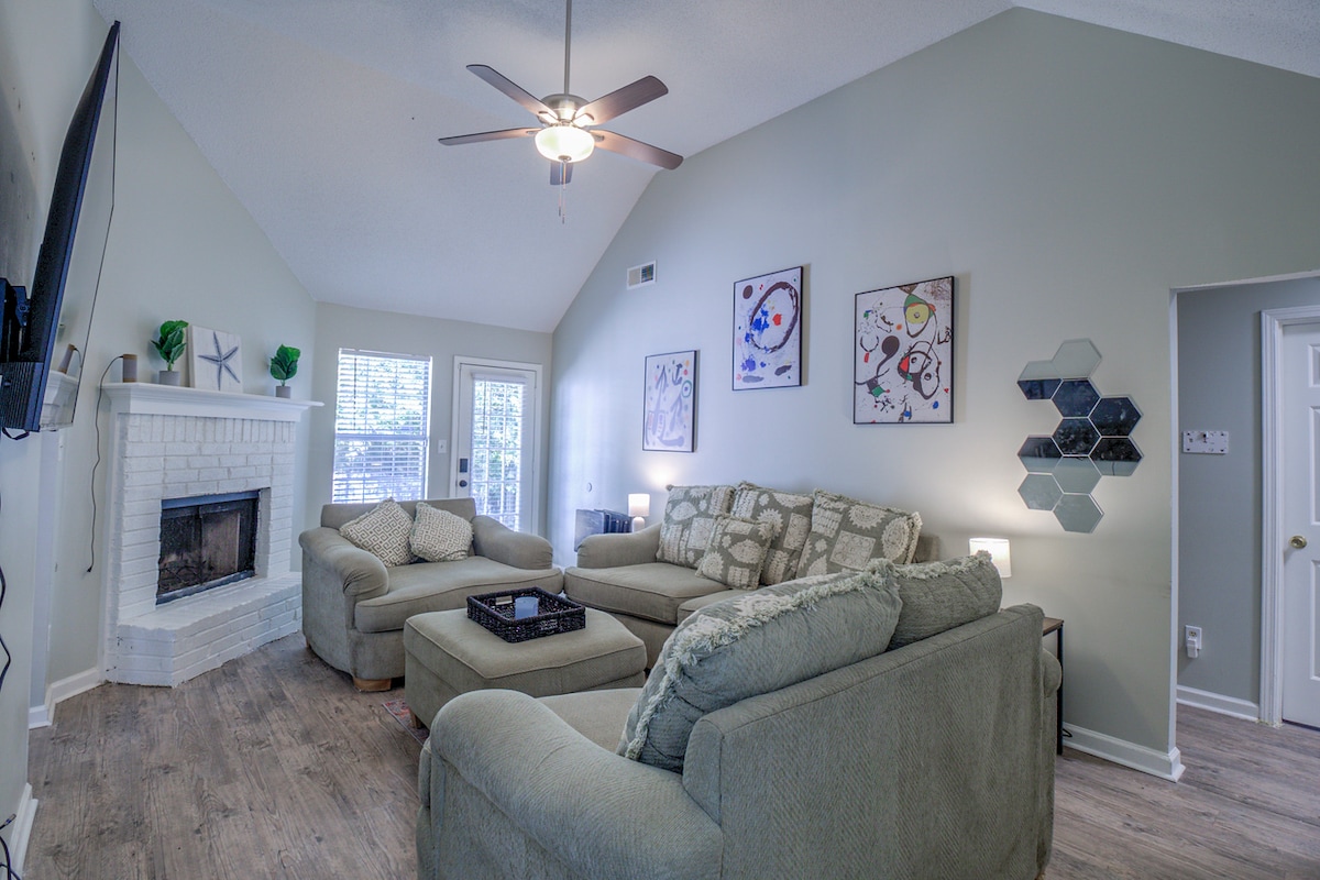 A spacious living area features a comfortable sectional sofa surrounding a central coffee table. Natural light enters through large windows, while a stylish ceiling fan enhances the room's airflow. A decorative fireplace and colorful wall art add a touch of charm.