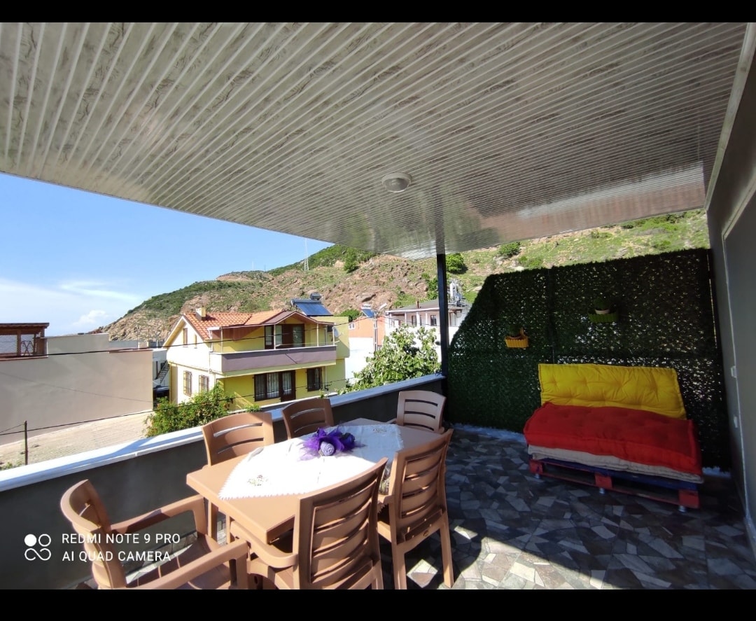 A spacious balcony features a large dining table surrounded by several chairs. A colorful outdoor couch adds comfort, with a lush green wall providing a serene backdrop. The view includes distant hills and nearby buildings, enhancing the open-air experience.