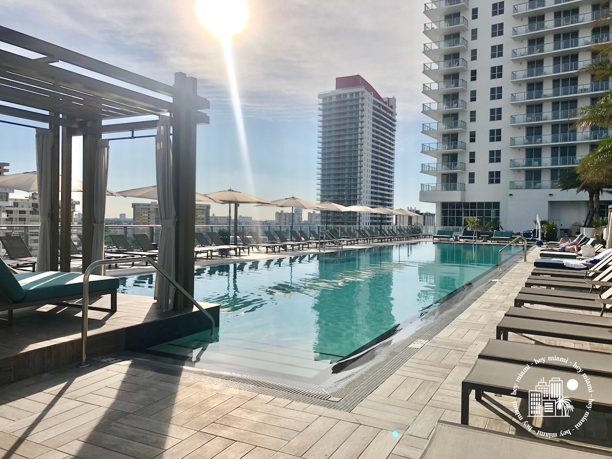 A large pool area is seen with lounge chairs positioned around the water. Umbrellas provide shade over some seating. The setting features tall buildings in the background and a bright sun reflecting off the pool's surface.