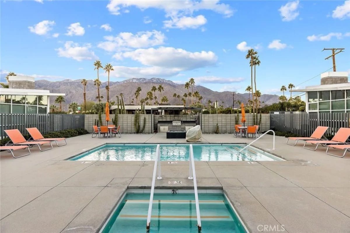 A clear view of a swimming pool surrounded by lounge chairs under a bright sky. Tall palm trees line the perimeter, with mountains visible in the background. A hot tub sits adjacent to the pool, providing a relaxing atmosphere. The area is well-maintained and inviting.