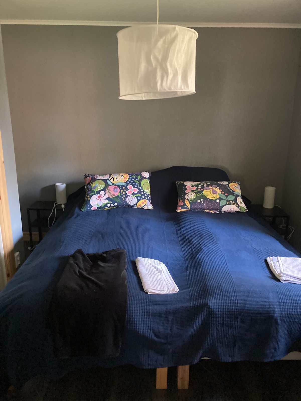 A cozy sleeping area features a spacious bed with a dark blue coverlet and colorful accent pillows. Two bedside tables are situated on either side, each with a lamp. Fresh towels are neatly arranged at the foot of the bed, creating a welcoming environment.