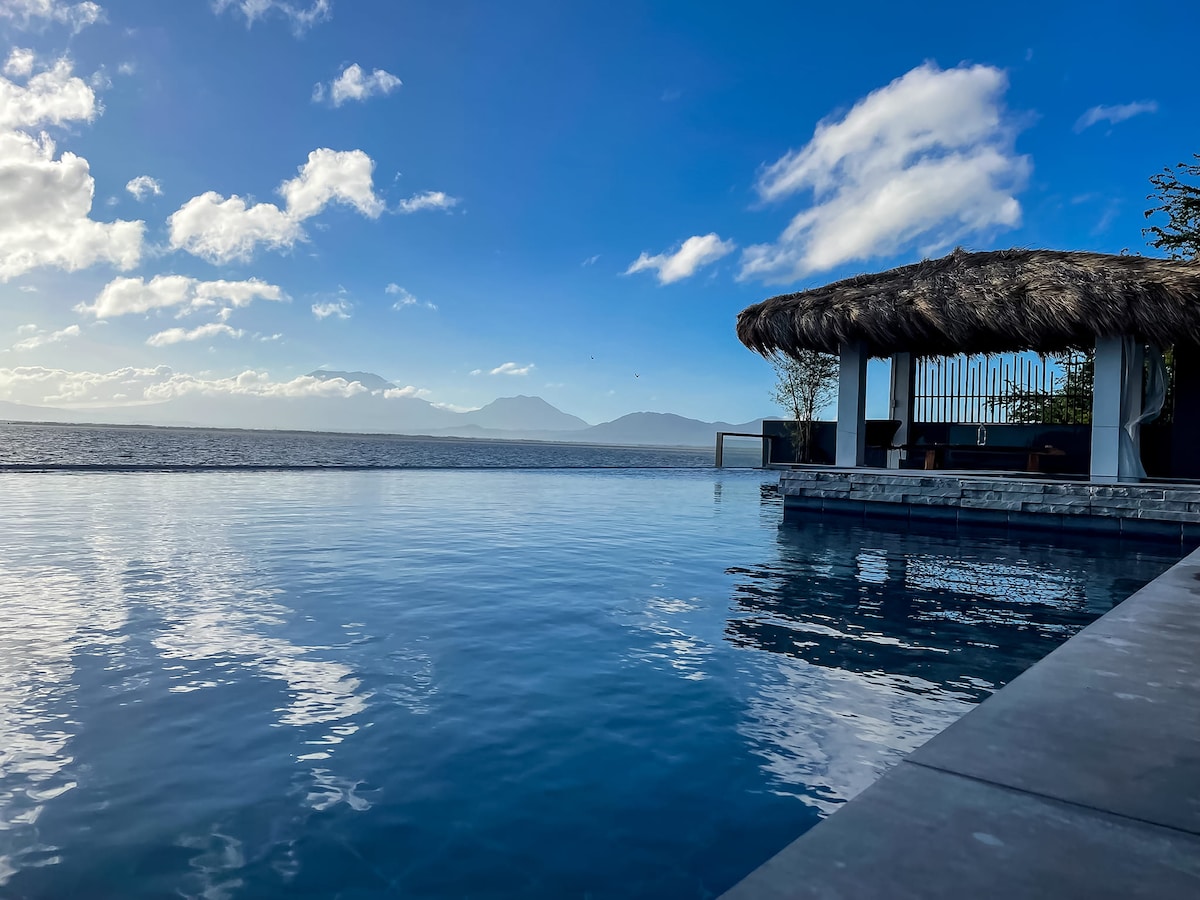A serene infinity pool is positioned alongside a thatched-roof bar, with expansive views of the lake and distant mountains. The water reflects the blue sky and fluffy white clouds, creating a peaceful atmosphere that invites relaxation.