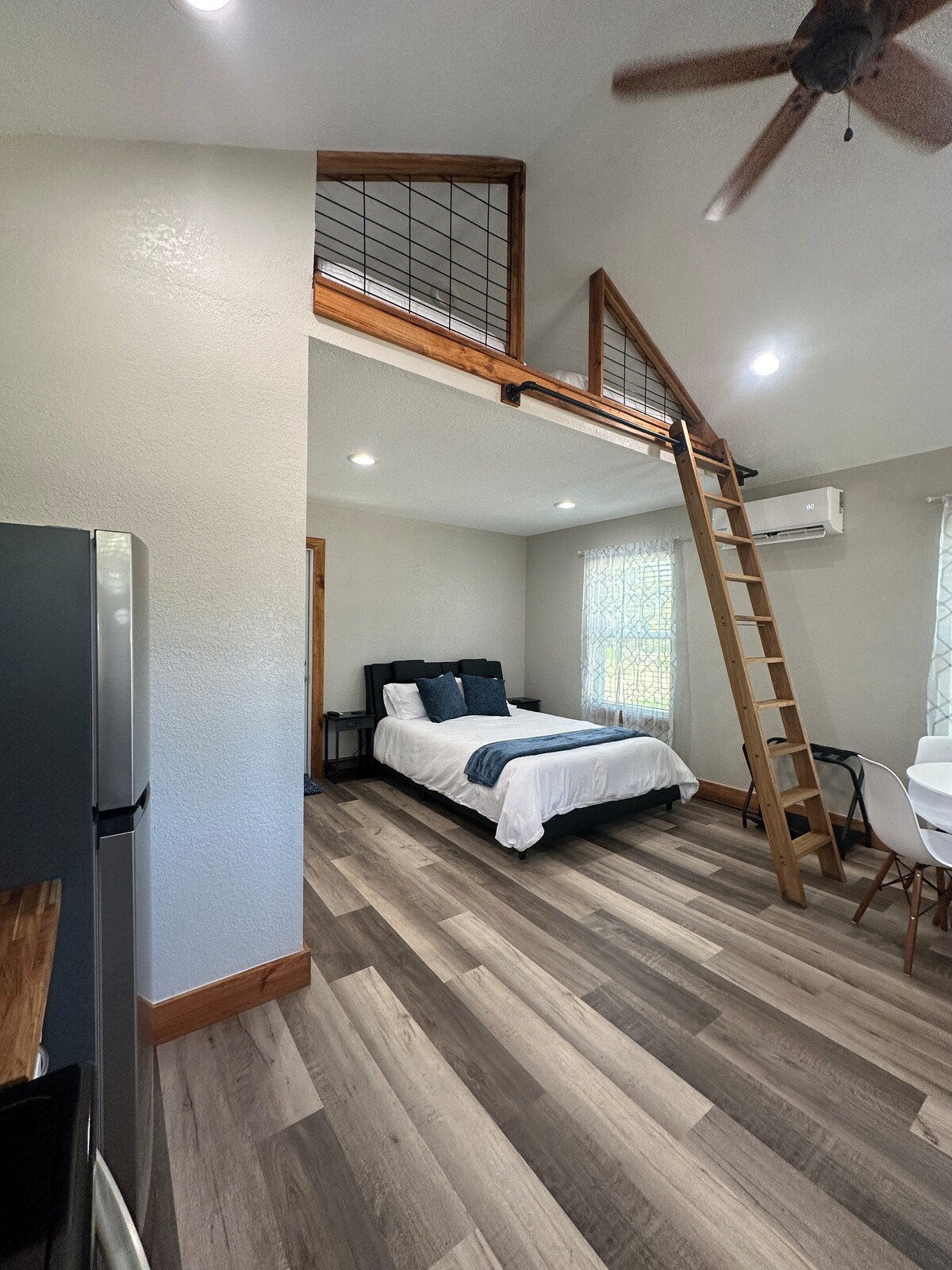 An open floor plan is showcased, featuring a queen bed with decorative pillows and a loft area accessible by a wooden ladder. A small dining set is visible in the background, along with a kitchenette that includes a fridge and modern finishes. Natural light enters through a window dressed with light curtains.