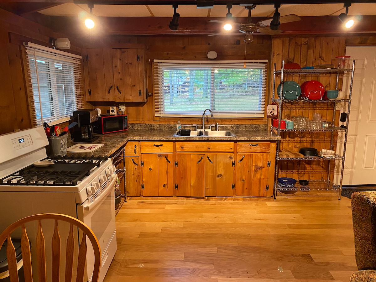 The kitchen features warm wooden cabinetry and a spacious countertop. A white stove and microwave are positioned next to a sink beneath a window, allowing natural light to enter. Colorful dishes and glassware are organized on a nearby open shelf.