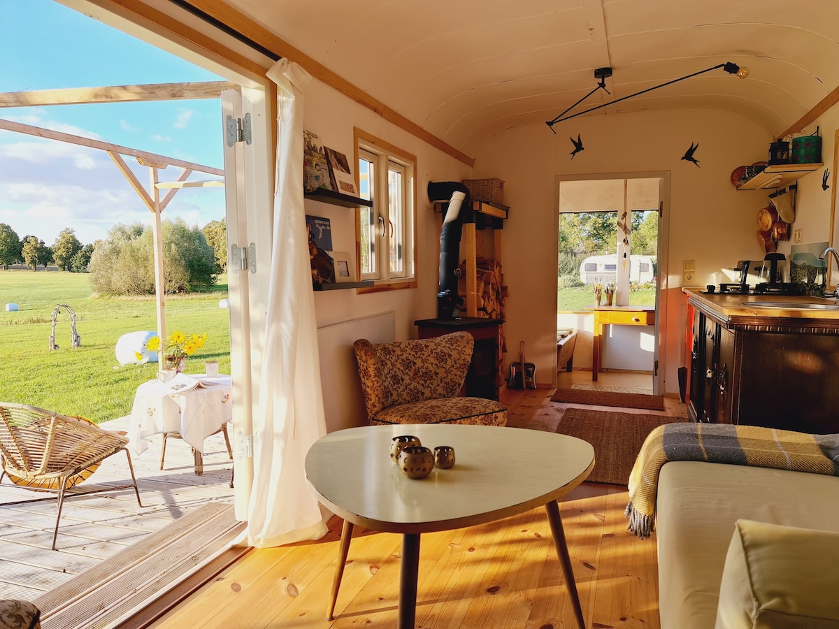 An inviting interior space features a cozy seating area with two patterned chairs and a small table. Natural light floods through the open French doors, leading to a wooden terrace. A warm wooden floor and tasteful decor create a serene atmosphere, suggesting functionality and comfort.