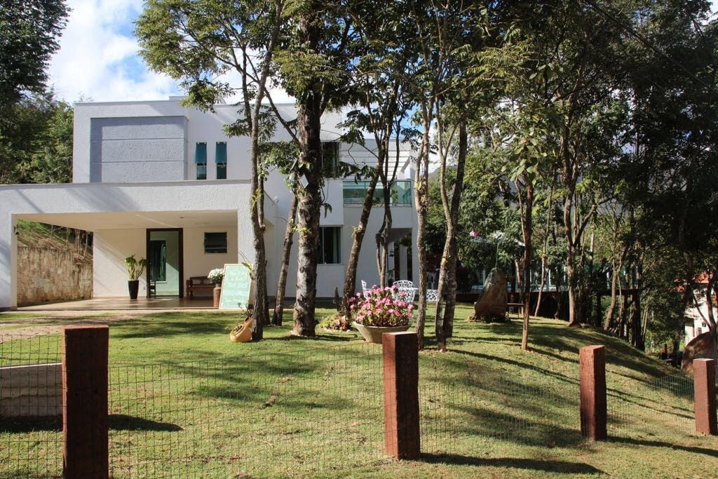 A modern two-story house is situated among greenery, featuring large windows that invite natural light. The building blends into the landscape, with a patio area in front and well-kept flower beds. Trees provide a sense of privacy and connection to nature.