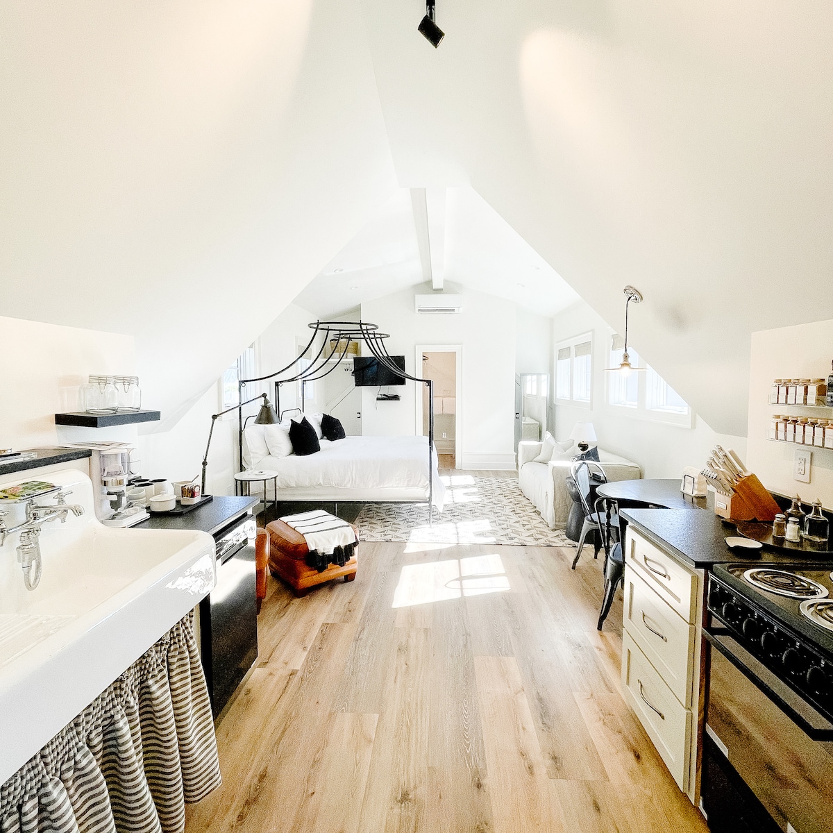 An open-concept studio apartment features a spacious layout with high ceilings. A four-poster bed with textured bedding is centered in the room, adjacent to a comfortable sitting area. A fully equipped kitchen includes modern appliances, while sunlight pours in through the large windows, enhancing the bright ambiance.