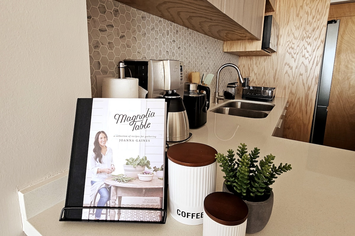 A well-organized kitchen area features a decorative cookbook displayed on a stand. Kitchen appliances, including a coffee maker and kettle, are arranged on the countertop, complemented by a small plant and containers labeled 'COFFEE.' Soft lighting highlights the modern design.