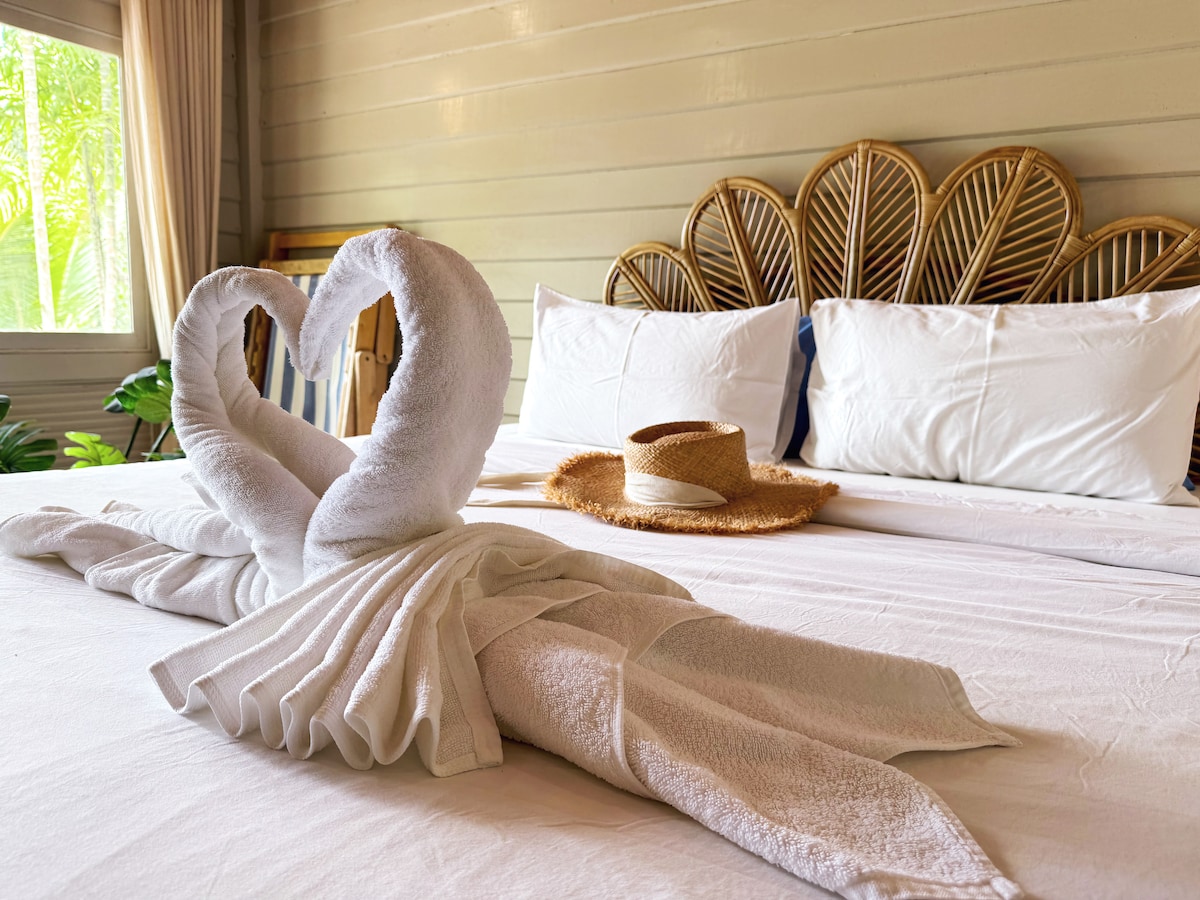 A neatly arranged bed features a large white linen spread and two decorative towel swans forming a heart shape. A straw hat is placed on the bed, with two potted plants visible near a window, allowing natural light to brighten the room.
