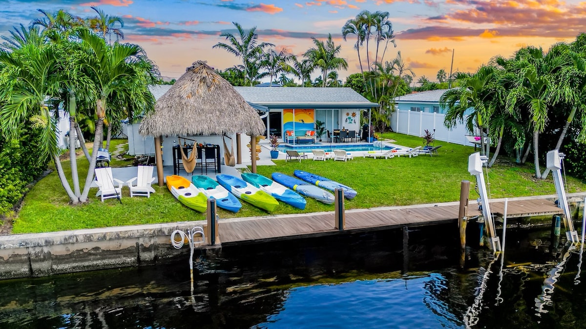A vibrant backyard features a thatched-roof tiki bar adjacent to a heated pool, surrounded by lush palm trees. Colorful kayaks line the grass near the water's edge, while a dock provides access to the canal. A serene sunset casts warm tones across the scene.