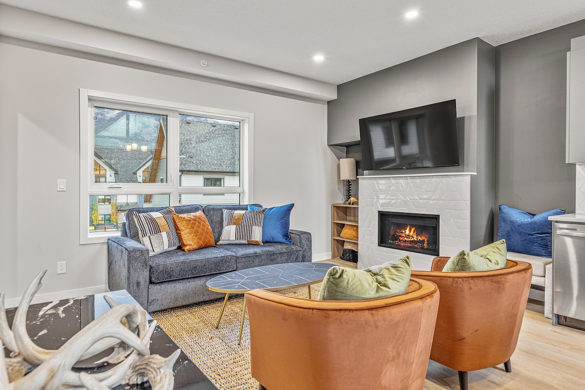 The living area features a comfortable sofa adorned with decorative pillows, a flat-screen TV mounted on the wall, and a fireplace that adds visual interest. Natural light is invited in through a large window, showcasing the surrounding architecture.