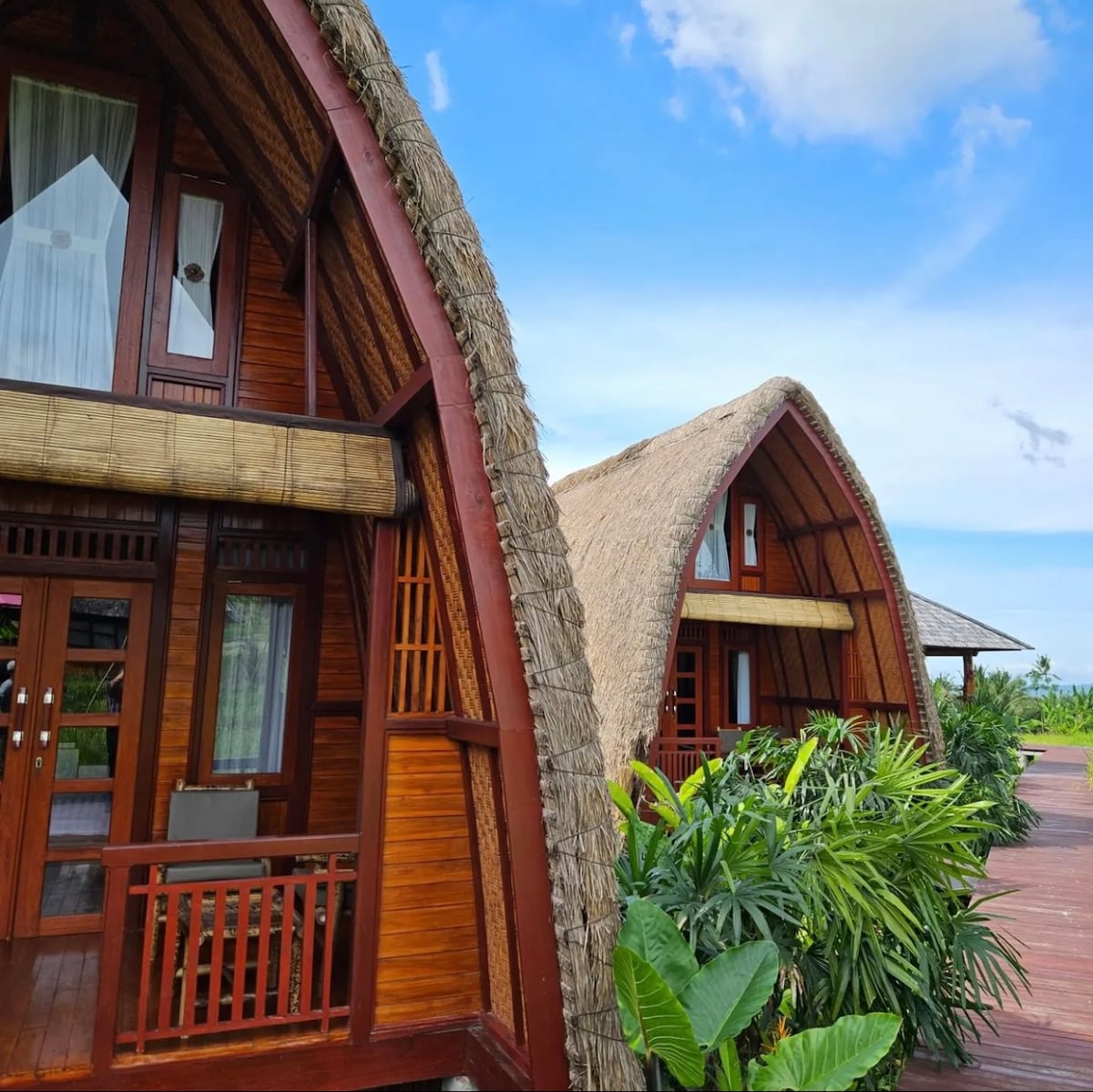 Two uniquely designed cottages are presented with curved roofs made of thatch, set against a bright sky. Surrounding greenery creates a natural border, and wooden details are visible on the façades. A wooden deck extends in front, offering a connection to the tranquil environment.