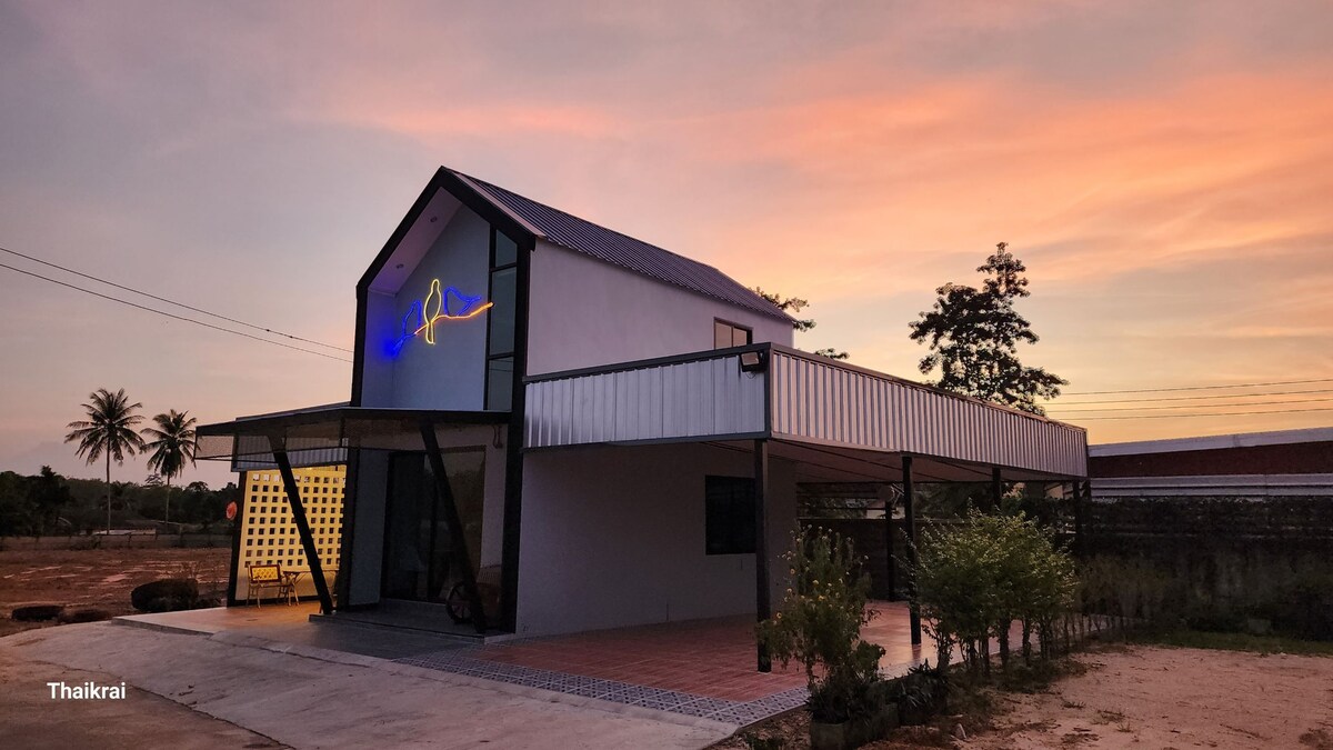 A modern structure with a unique design is set against a colorful sunset sky. The building features large windows and a distinct roof, while the entrance is framed by a welcoming porch and landscaped greenery.