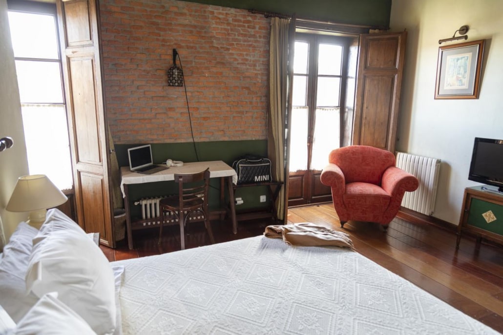 A spacious room features a large bed adorned with white linens. A wooden desk is positioned by a window, accompanied by a wooden chair. A red armchair adds a pop of color, while vintage wooden shutters and a framed art piece can be seen on the walls.